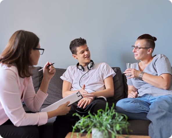 Therapist in pink interviewing two people on a couch. They appear to be a couple; one has a drink, the other, headphones.
