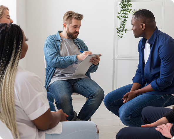 Group therapy session: A therapist with a clipboard listens to a man while others sit in a circle. Natural light fills the room.