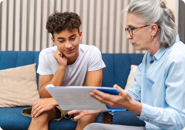 A therapist points at paperwork, speaking to a teenage boy on a blue couch.