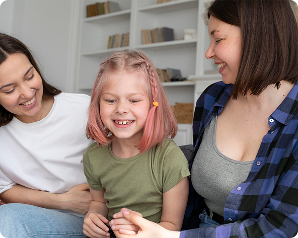Two women and a young girl with pink hair sit closely together, smiling and laughing. They are indoors.