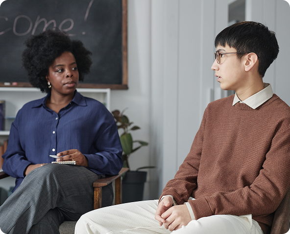 A therapist sits across from a young man, both in chairs, engaged in a counseling session. The woman holds a notepad, and the man appears to be talking.