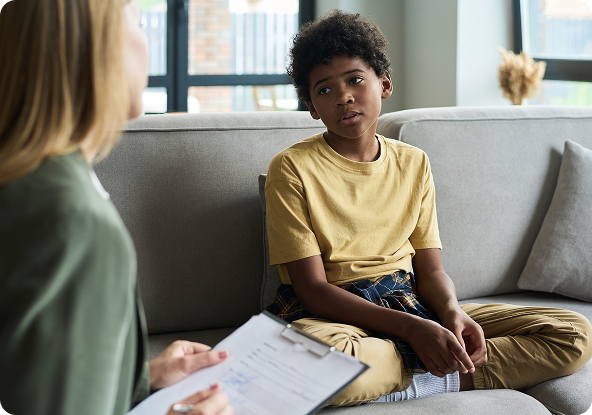 A young Black boy sits on a couch looking concerned while facing a therapist holding a clipboard.