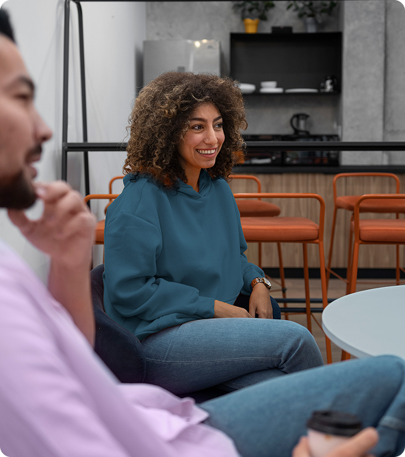 Woman with curly hair smiles while talking to a man. They are seated in a modern cafe setting.