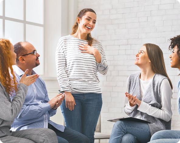 A smiling woman in a group therapy session, gesturing towards herself. Other members clap and smile in a bright room.