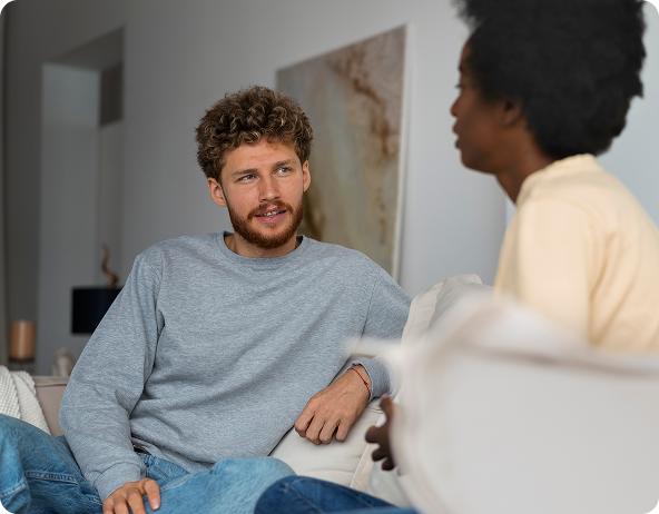 A man with curly hair and a beard talking to a Black woman while seated on a couch. They are indoors.