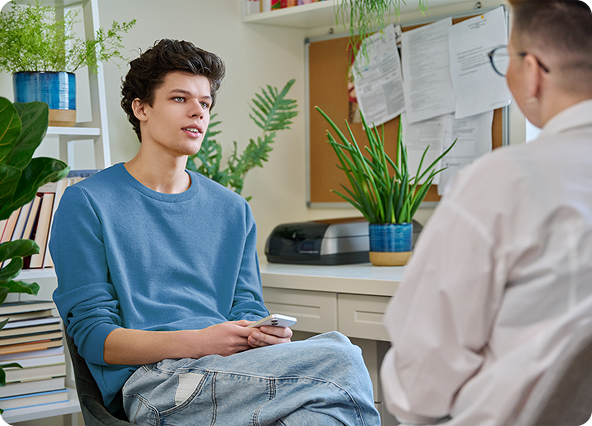 A young man in a blue sweater talks to a person in a white shirt, likely a therapist, in a home office setting.