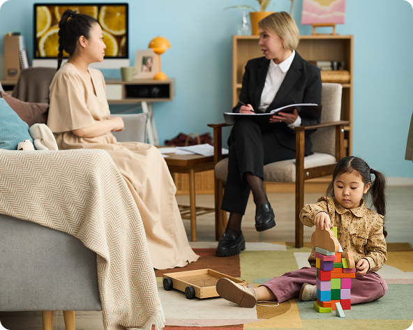 A woman and a therapist in a counseling session, while a child plays nearby with blocks.