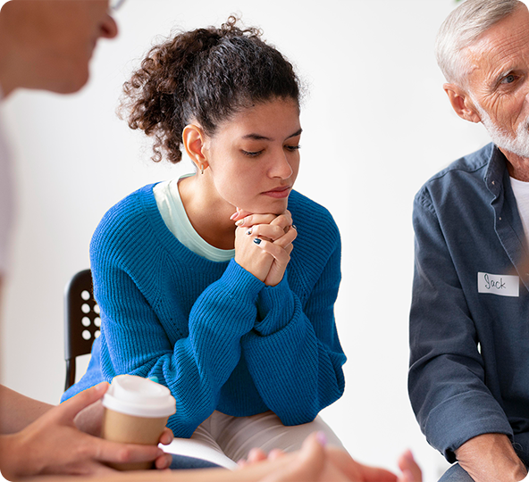 A young woman in a blue sweater sits in a circle, looking down with a pensive expression. She’s in a group, possibly a support session.