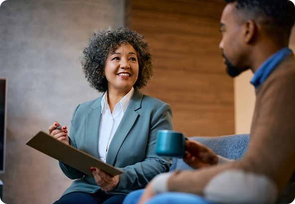 A smiling woman in a blazer holds a clipboard, talking to a man holding a mug in a casual setting.