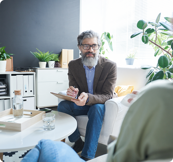 Therapist with a beard and glasses taking notes during a session with a patient in a well-lit office.