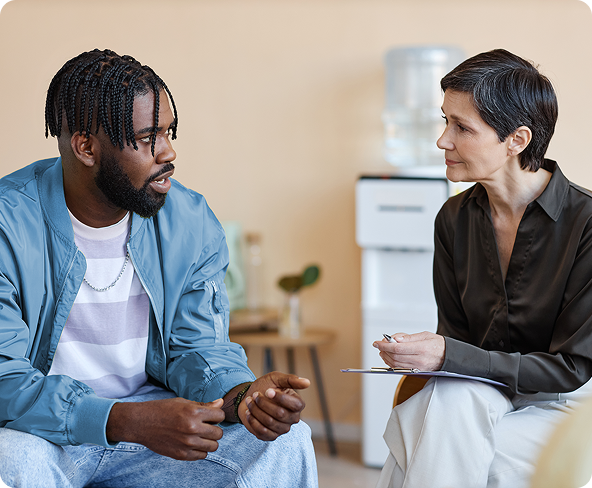 A Black man and a white woman in a therapy session. The man gestures while speaking; the woman listens, holding a notepad.