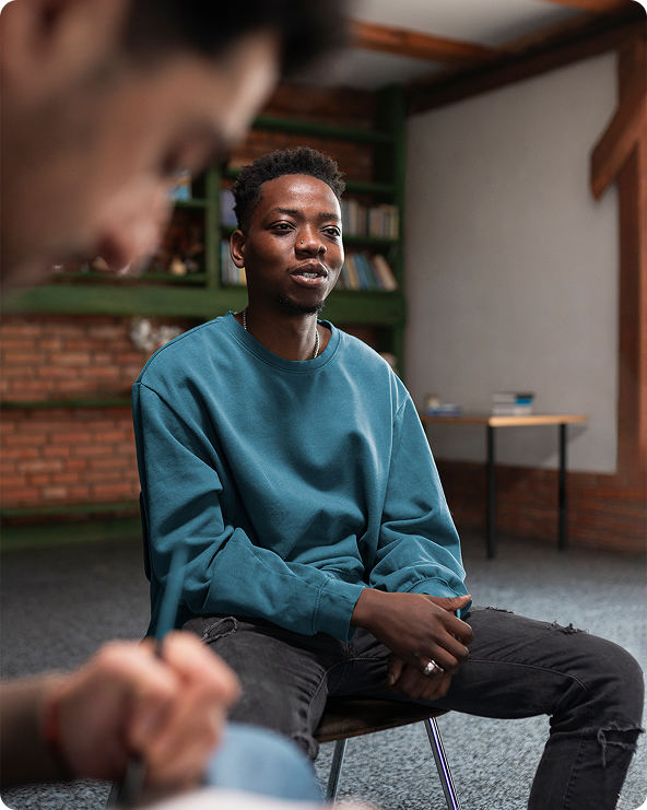 A young Black man sits in a chair and talks to someone off-camera. He wears a teal sweater and ripped jeans.