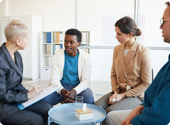 A group therapy session with a therapist and three patients seated in a circle; the therapist is taking notes.