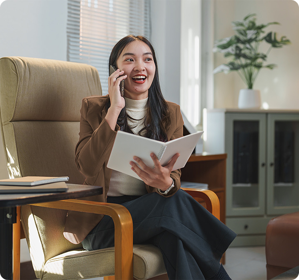 Asian woman in a brown blazer sits, smiling, talking on the phone, holding a notepad, in a well-lit office.