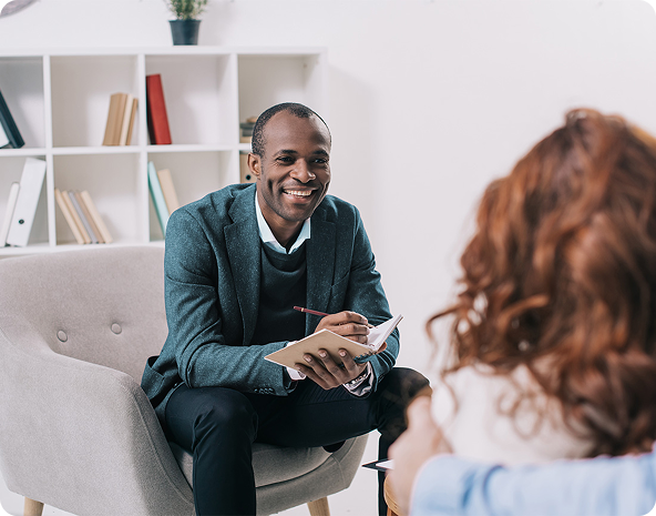 A Black therapist smiles while taking notes, facing a patient with red hair seated in front of him in an office setting.