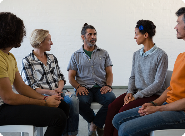A diverse group in a circle, engaged in a conversation, possibly a therapy session. They are seated in white chairs against a plain white wall.