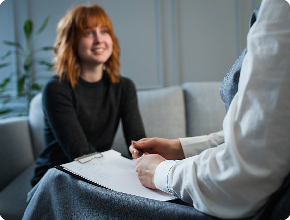 A person with red hair smiles while talking to someone holding a clipboard in a counseling session setting.