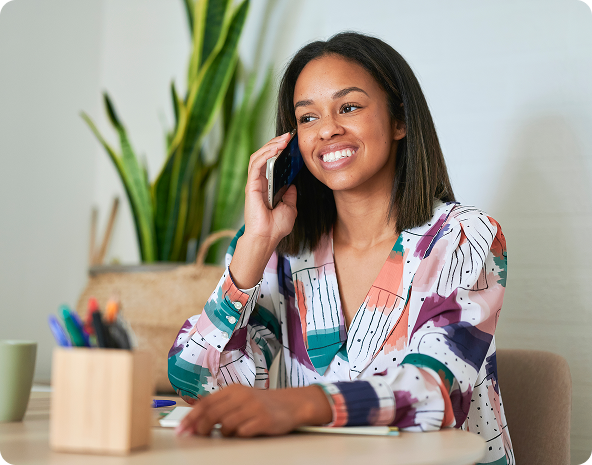 Young woman with dark hair smiling, talking on a cell phone at a desk. A plant and pens are in the background.