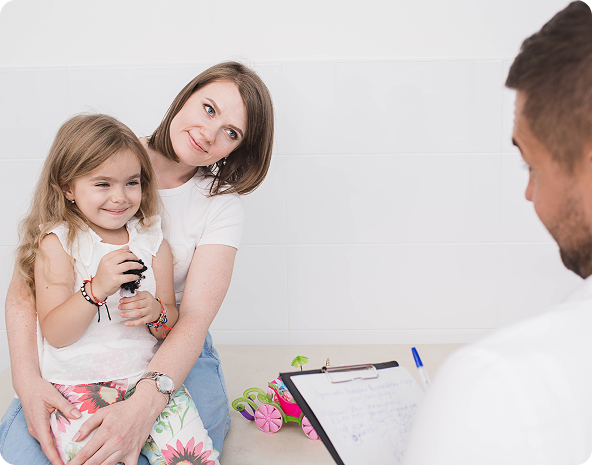Mother holding a young girl at a doctor's appointment. They smile at the doctor who is holding a clipboard.