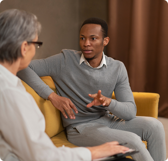 A Black man talks to a person with gray hair in a counseling session, both seated on a yellow couch.