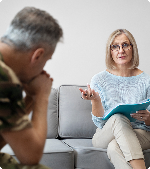 A therapist in glasses gestures while speaking to a soldier in uniform, both seated on a grey couch.