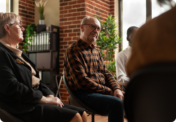 Group therapy session with diverse people in chairs, brick wall background. Older man with plaid shirt appears thoughtful.