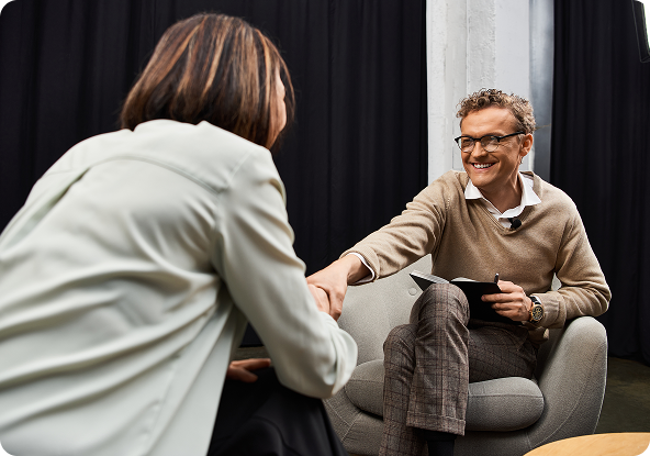 A woman and a man shake hands in an interview setting. The man smiles, holding a tablet; the woman is seen from behind.