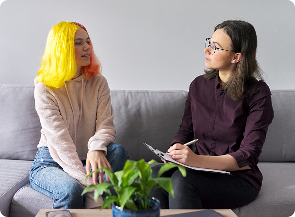 Teenager with colorful hair talking to a therapist. They are sitting on a couch in an office setting.