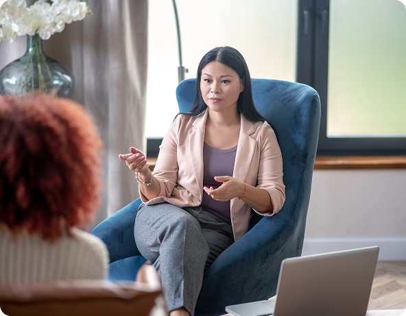 A therapist talking to a patient in a cozy office setting. The therapist, a woman in a light pink blazer, gestures while seated in a blue armchair.