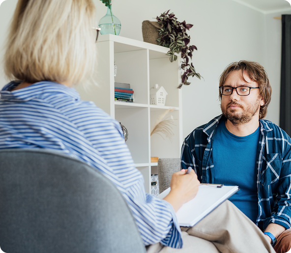 Therapist with blonde hair and client in glasses having a counseling session indoors; the therapist holds a clipboard.