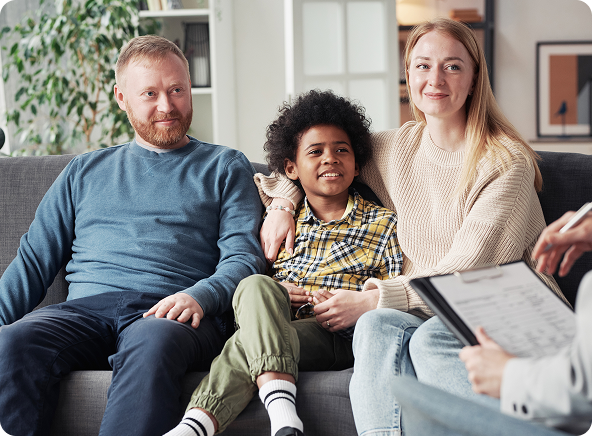 A family of three, a white man and woman with a Black child, sit on a couch smiling while consulting with a therapist.