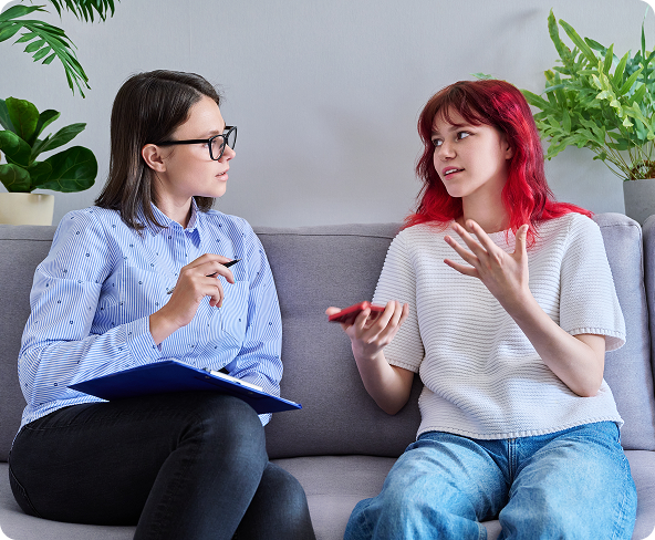 A therapist listens attentively to a young woman with red hair. They are seated on a couch, the therapist taking notes.