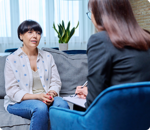 A woman is talking to a therapist. The therapist sits in a blue chair, taking notes. They are indoors, on a couch.