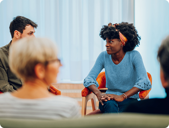 A diverse group sits in a circle, likely in a therapy session. A Black woman in blue speaks, looking at another person.