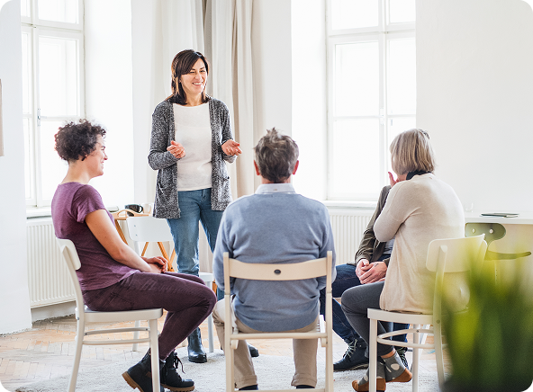 A woman leading a support group, holding papers and speaking to four seated people in a bright room.