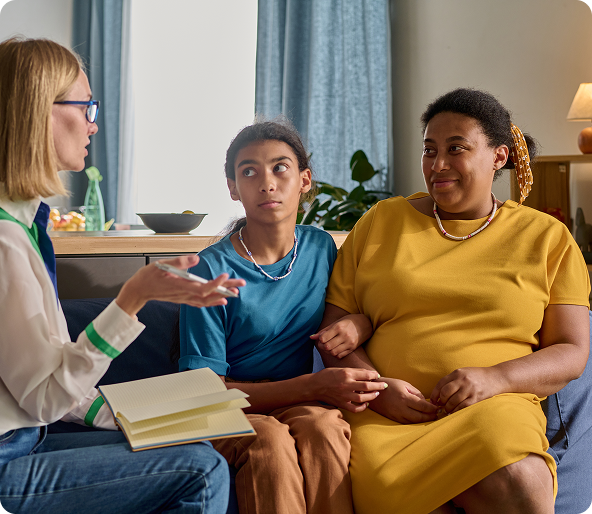 A therapist in glasses gestures while speaking to a teen and her mother, who are sitting on a couch. The teen looks concerned.