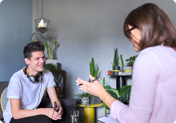 A young man smiles while talking to a woman in a therapy session. They are sitting in a room with plants and a lamp.