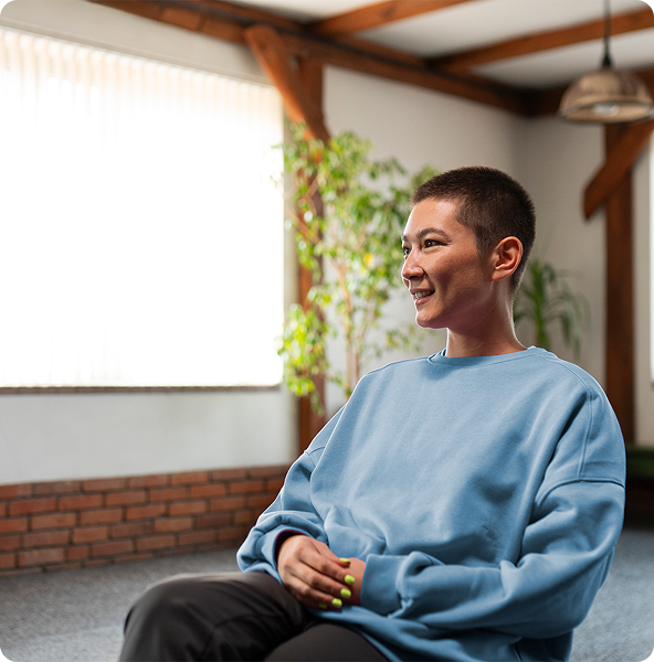 Woman with short hair smiles, seated in a bright room. She wears a blue sweatshirt and has her hands clasped.