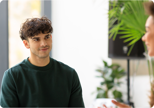Man in green sweater smiles at someone off-screen in a bright room with plants.