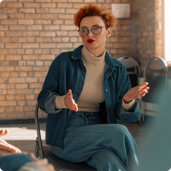 Woman with red curly hair, glasses, and a denim jacket gestures during a group discussion in a room with exposed brick walls.
