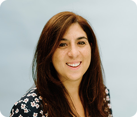 Woman with long brown hair smiles at the camera, wearing a black floral print top, with a light blue background.