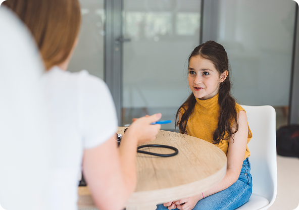 Woman holding a pen talks to a young girl sitting at a round table in a bright room; girl smiles.