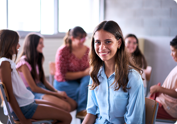 Smiling young woman in a light blue shirt; group of young people seated in chairs in the background.