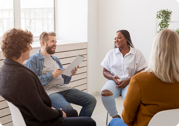 Group therapy session with four people in a bright room. They sit in a circle, smiling and engaged in conversation.