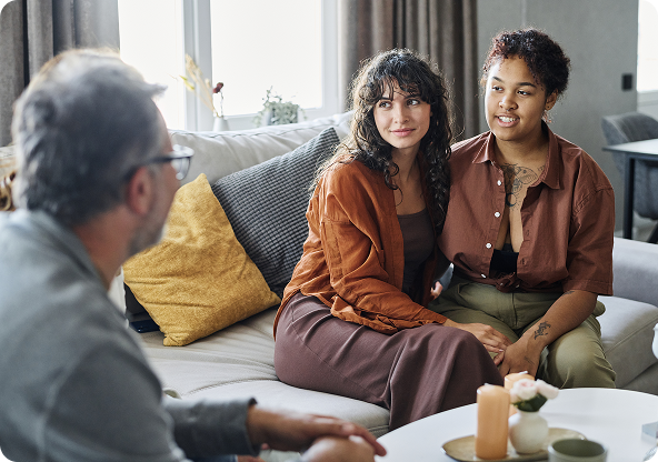 A counselor talks with a smiling couple on a sofa in a living room. The women have dark hair, and one has arm tattoos.