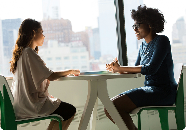 Two women sit at a white table by a window. One woman gestures while speaking, the other listens attentively.
