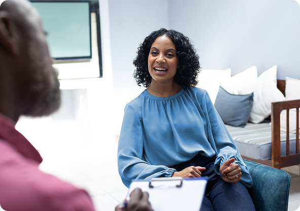 Woman in blue top smiles and gestures while speaking to a person holding a clipboard in a counseling room.