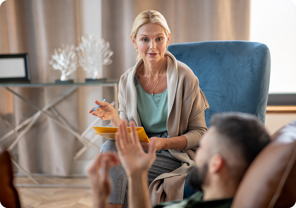 A blonde therapist gestures while speaking to a man reclining on a couch in a neutral-toned office.