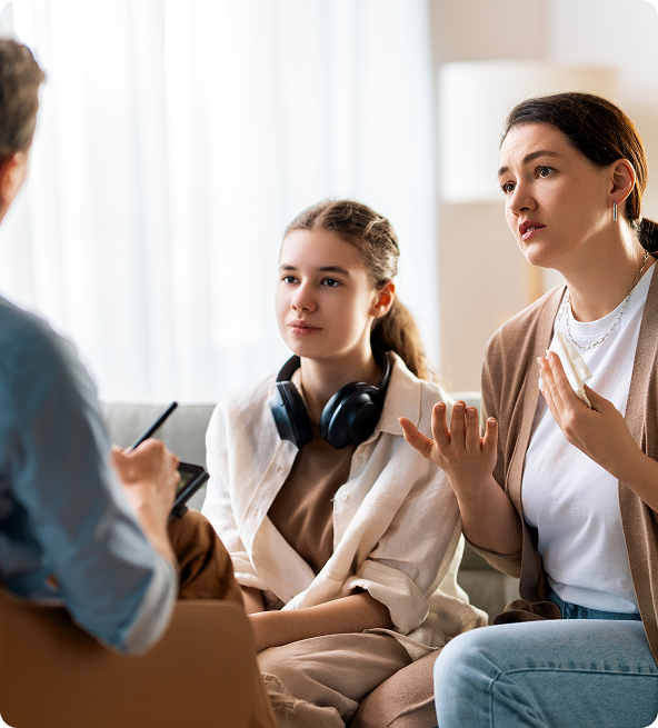 A therapist talking with a mother and teenage daughter in a brightly lit room. The mother gestures while speaking.