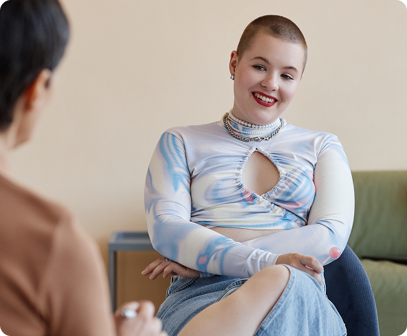 A person with a shaved head smiles and sits in a chair, facing another person, in a room with a light-colored wall.